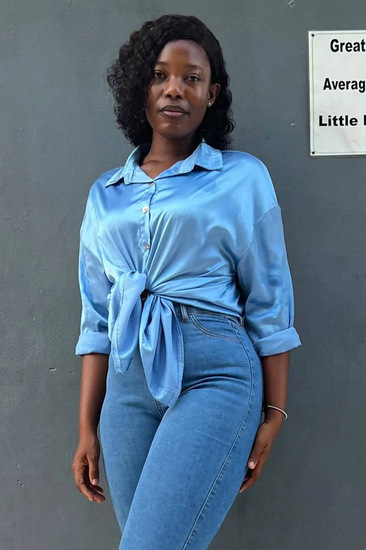 After. Wuenam Glory before and after: in a cream floral-print blouse and jeans inside a clothing store, and in a blue satin blouse tied at the waist with jeans, against a plain wall.